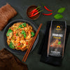 Plated dish of noodles with shrimp and greens, accompanied by a package of SIAM Brown Rice Vermicelli on a dark surface.