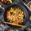 Noodles with vegetables in a pan with chopsticks, surrounded by ingredients and a brand package.